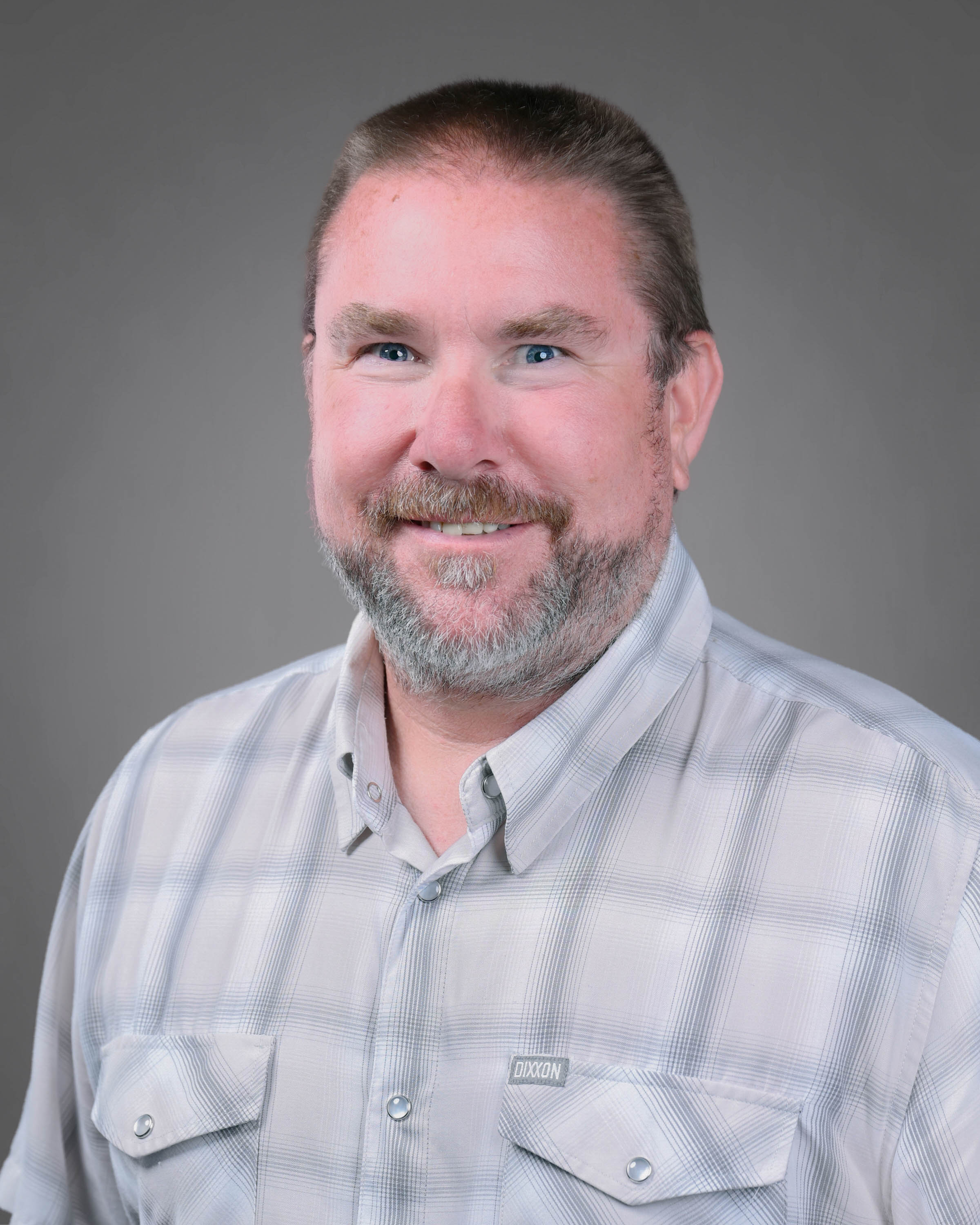 Smiling man with short hair and a beard wearing a plaid collared shirt.